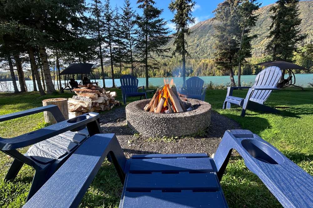 A cozy lakeside fire pit with Adirondack chairs, surrounded by lush greenery and mountains in Cooper Landing, Alaska.