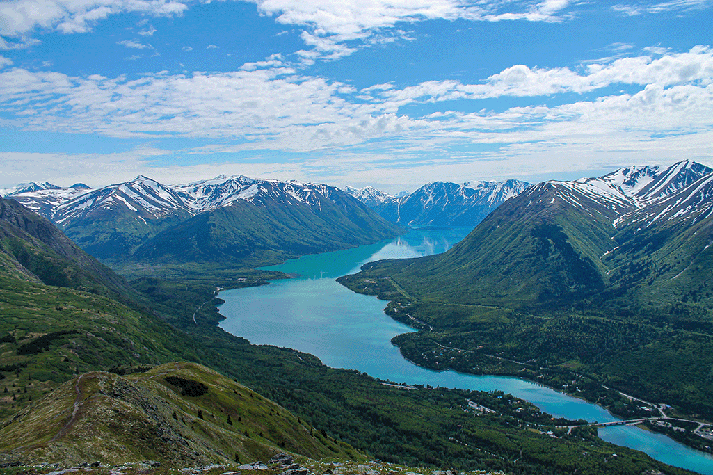A scenic view of a turquoise lake winding through lush green valleys, surrounded by snow-capped mountains under a blue sky.